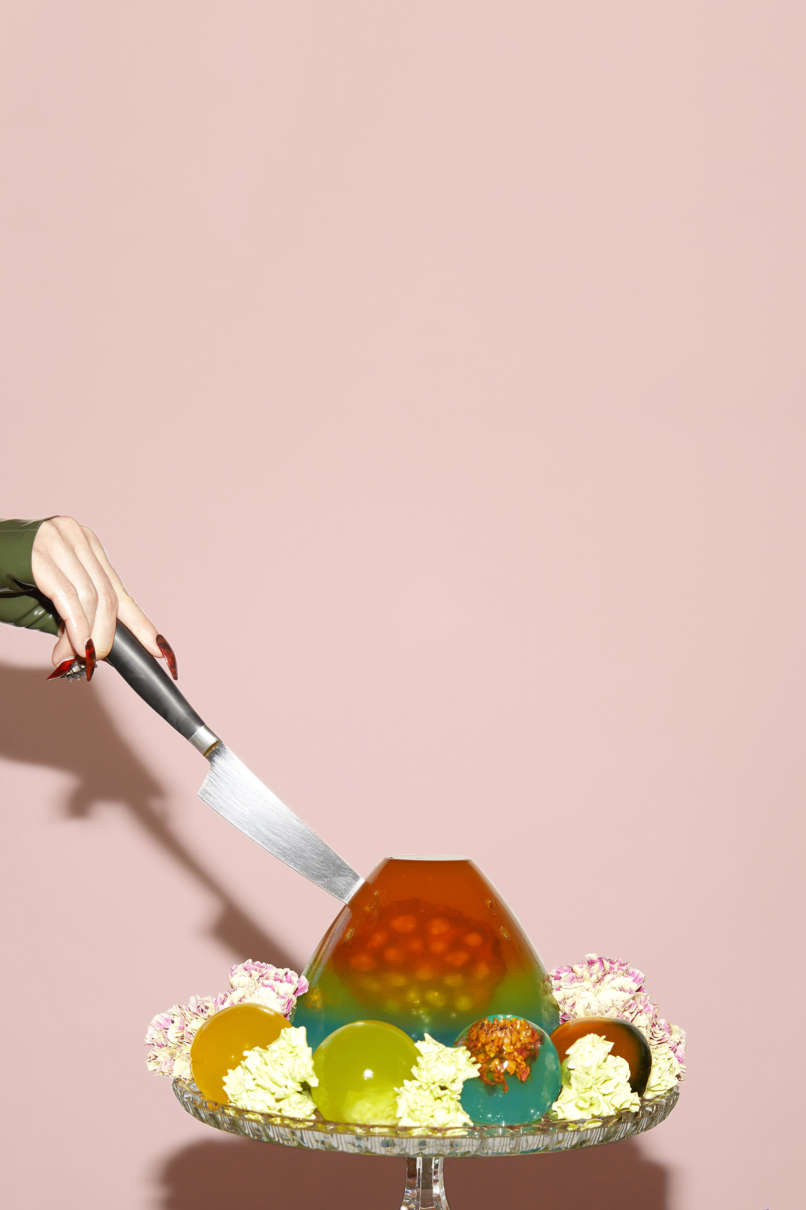 A rainbow jello cake on a pink background being cut with a knife